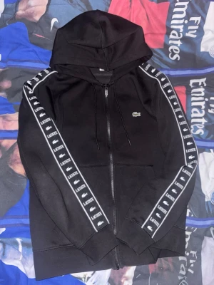 Svart Lacoste zip hoodie - Svart hoodie från Lacoste med dragkedja framtill och huva med snören. Ärmarna har vita ränder med Lacoste-logga längs hela sidan. Klassisk krokodil-logga på bröstet. Perfekt för en sportig och stilren look. Priset går alltid att diskutera m!