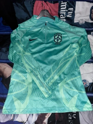 Grön Brasilien träningströja Nike - Snygg grön långärmad träningströja från Nike med Brasilien-emblem på bröstet. Tröjan har halv dragkedja framtill och ett grafiskt mönster i ljusgrönt över hela plagget. Perfekt för fotbollsträning eller uppvärmning. Priset går alltid att diskuteras!