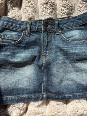 Blå jeanskjol i klassisk modell - Säljer en blå jeanskjol med klassisk femficksdesign och slitna detaljer. Kjolen har knapp och dragkedja framtill samt kontrastsömmar. Perfekt för dig som gillar en enkel och tidlös denimlook.