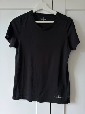 Svart tränings-tshirt från Ronhill - Säljer en svart tränings-tshirt från Ronhill i storlek S. T-shirten har rund halsringning, korta ärmar och är tillverkad i ett lätt och snabbtorkande material som passar perfekt till löpning eller gym. Diskret logga nere vid fållen.