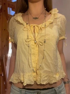 Gul blus med volang och knytband - Söt gul blus med korta ärmar, volangkant och broderade detaljer. Blusen har två knytband framtill och en krage med spets. Storlek M men jag skulle säga att den passar som S.