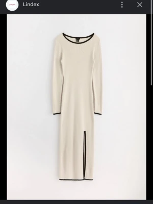 Långärmad beige klänning med slits - Stilren beige långklänning från Lindex med svarta kontrastkanter vid hals, ärmslut och nederkant. Klänningen är ribbad, har lång ärm och en snygg slits framtill för extra edge. Perfekt för dig som gillar minimalistisk och modern stil.