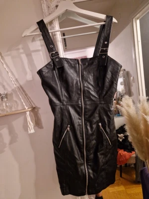 Svart kort klänning i skinnimitation - Snygg svart kort klänning i skinnimitation med breda axelband och spännen. Dragkedja framtill och två snedställda fickor med dragkedjor. Perfekt för en edgy look och lätt att styla med boots eller jacka. Aldrig använd, bara hängt i garderoben. Storlek 38