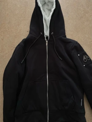 Moose Knuckles Bunny - Säljer en svart hoodie från Moose Knuckles med vit fluffig foder i huvan. Hoodien har dragkedja framtill, två stora fickor och en patch med logga på ärmen. Perfekt för dig som vill ha något både snyggt och bekvämt. Materialet är mjukt och värmande. Nyskick!