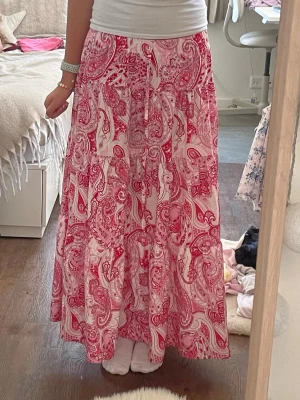 Rosa och vit långkjol med paisleymönster - Säljer en flowy långkjol i vitt och rosa med snyggt paisleymönster. Kjolen har en avslappnad passform och är perfekt för varma dagar. Den är lätt och luftig, vilket gör den superbekväm att bära.