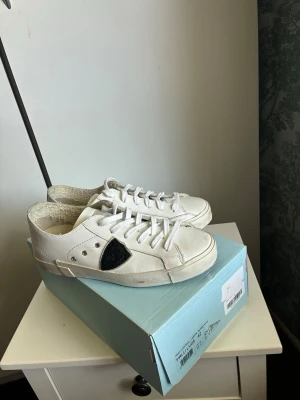 Vita låga sneakers med blåa skosnören - Säljer ett par vita låga sneakers i läder med svart detalj på sidan. Skorna har både vita och blåa skosnören som kan bytas ut efter stil. Klassisk design med rund tå och platt sula, perfekta för en avslappnad look.