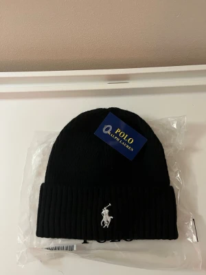 Svart mössa från Polo Ralph Lauren - Snygg svart ribbstickad mössa från Polo Ralph Lauren med klassisk broderad logga i vitt framtill. Mössan har uppvikt kant och är tillverkad i mjukt material som håller dig varm. Perfekt accessoar för kyliga dagar.