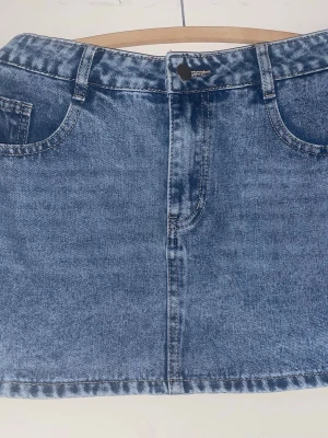 Blå jeanskjol med fickor - Säljer en klassisk blå jeanskjol i kort modell med fem fickor och bälteshällor. Kjolen har en rak passform och stängs med knapp och dragkedja framtill. Perfekt till sneakers eller boots för en avslappnad look.