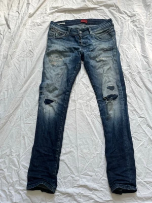 Slim fit jeans från Jack & Jones - Snygga blå slim fit jeans från Jack & Jones, modell Glenn. Jeansen har slitningar och hål på båda benen för en trendig, avslappnad look. Klassisk femficksmodell med knappgylf och ljusa tvättade partier. Perfekta för en cool streetstyle.