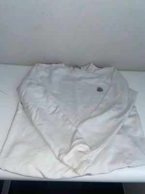 Vit sweatshirt från Moncler - Snygg vit sweatshirt från Moncler med rund halsringning och broderad Moncler-logga på bröstet. Tröjan har långa ärmar och en klassisk, stilren design som passar perfekt till jeans eller joggers.