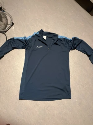Marinblå Nike Dri-FIT träningströja - Snygg marinblå långärmad träningströja från Nike med ljusblå detaljer på axlarna. Tröjan har en halv dragkedja framtill och är tillverkad i Dri-FIT-material som andas. Perfekt för träning eller uppvärmning eller nått man kan gå ut med när man hänger med vänner 