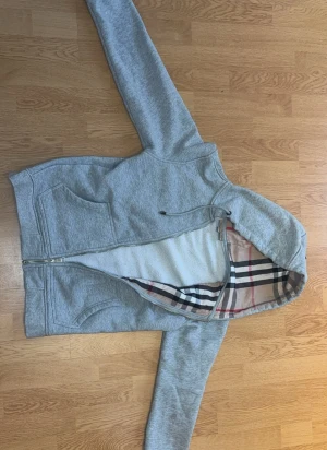 Burberry Hoddie - Snygg grå hoodie med dragkedja och huva. Insidan av huvan har Burberrys ikoniska rutmönster i beige, svart, vitt och rött. Mjuk och skön bomullsblandning, perfekt för en avslappnad stil. Praktiska fickor framtill och justerbar huva.