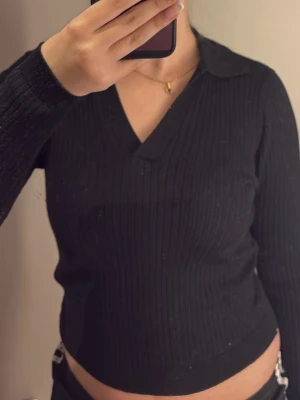 Svart ribbad långärmad polotopp - Säljer en svart ribbad långärmad topp med krage och v-ringning. Toppen har en slim passform och är perfekt för att skapa en clean och stilren look. Materialet känns mjukt och stretchigt, vilket gör den bekväm att bära hela dagen.