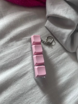 Nyckelring med rosa tangentbordsknappar - Säljer en unik nyckelring med fyra rosa tangentbordsknappar monterade på en transparent plastbas. Perfekt accessoar för dig som gillar datorer eller vill ha något annorlunda på nyckelknippan.