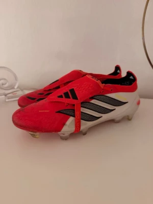 Röda Adidas Predator fotbollsskor - Säljer ett par snygga Adidas Predator fotbollsskor i rött, vitt och svart. Skorna har klassiska tre ränder på sidan, låg profil och snörning med täckande rem. Yttermaterialet är syntet och sulan har fasta dobbar för bra grepp på planen.