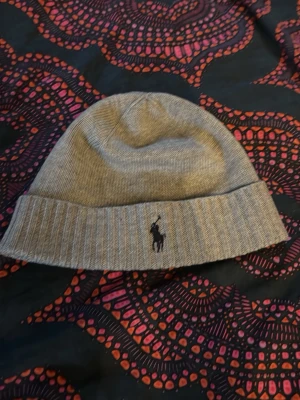 Grå mössa från Polo Ralph Lauren - Säljer en stilren grå mössa från Polo Ralph Lauren med klassisk ribbad kant och broderad logga framtill. Mössan är stickad i mjukt material och passar perfekt för kalla dagar.