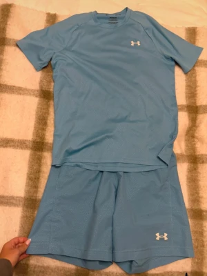 Blått träningsset från Under Armour - Säljer ett blått träningsset från Under Armour med t-shirt och shorts. Setet är gjort i ett lätt och snabbtorkande funktionsmaterial, perfekt för träning eller löpning. Både tröjan och shortsen har Under Armour-logga i vitt.