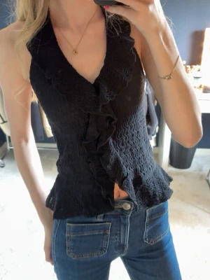 Svart topp med volang - Använd 1 gång!!!Säljer en svart omlott topp med volangdetaljer och halterneck. Toppen har en vågig struktur och är figurnära i modellen. Perfekt för dig som vill ha en trendig och snygg topp till jeans eller kjol.