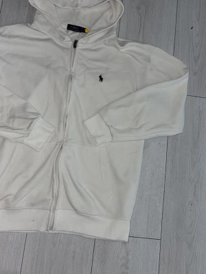 Vit hoodie från Polo Ralph Lauren - Säljer en vit hoodie från Polo Ralph Lauren med dragkedja framtill och klassisk logga broderad på bröstet. Hoodien har fickor på sidorna, ribbade muddar och en skön huva. Perfekt för en clean och stilren look.