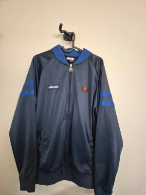 Blå tröja från Ellesse - Snygg marinblå tröja från Ellesse med blå detaljer på ärmarna och dragkedja framtill. Tröjan har ribbade muddar, hög krage och klassisk Ellesse-logga på bröstet. Perfekt för dig som gillar sportig och retro stil.