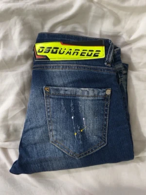 Blå Dsquared2 skinny jeans - Säljer ett par blå skinny jeans från Dsquared2 med slitningar, färgstänk och en stor gul logga på bakfickan. Jeansen har en trendig tvätt och smal passform som ger en cool streetwear-look. Perfekta för dig som vill sticka ut med din stil.