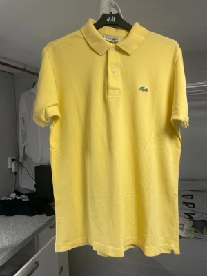 Gul pikétröja från Lacoste - Snygg gul pikétröja från Lacoste med klassisk krage och knappar framtill. Tröjan har korta ärmar och är tillverkad i bomull. Den ikoniska gröna krokodilloggan är broderad på bröstet. Perfekt för en fräsch och sportig look.         Storlek m-s