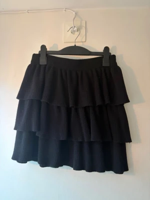 Svart volangkjol  - Säljer en svart kjol som är ribbad. Kjolen har en elastisk midja och kort modell.