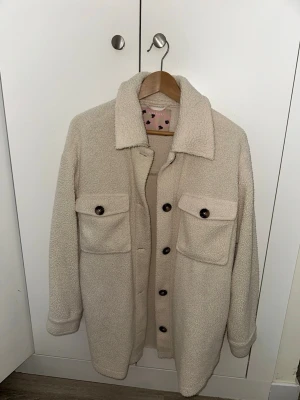 Beige teddy overshirt med fickor - Säljer en beige overshirt i mjukt teddy-material. Skjortan har två stora bröstfickor med knappar, klassisk krage och knäppning framtill. Perfekt för lager-på-lager och ger en avslappnad men trendig look.