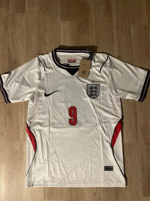 England fotbollströja Kane 9 - Säljer en vit England fotbollströja med svarta och röda detaljer. Tröjan har nummer 9 och namnet Kane på ryggen samt Englands landslagsemblem på bröstet. Korta ärmar och sportig passform, perfekt för match eller träning.