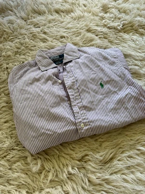 Lila och vit randig skjorta Polo Ralph Lauren s - Snygg skjorta från Polo Ralph Lauren i lila och vitt randigt mönster. Klassisk krage, knäppning framtill och broderad grön logga på bröstet. Perfekt för en stilren look.