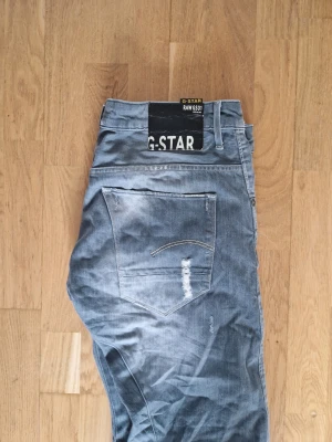 Grå jeans från G-Star RAW - Säljer ett par grå jeans från G-Star RAW med slitna detaljer och snygg tvätt. Jeansen har raka ben, klassiska bakfickor och tydlig logga på linningen. Perfekta för en avslappnad streetstil. Materialet är bomull. Storlek: W: 34, L:32.  Köpta från vinted. 
