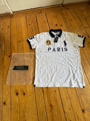 Polo Ralph Lauren - Paris - Vit pikétröja från Polo Ralph Lauren med mörkblå detaljer, broderad logga och texten PARIS på bröstet. Tröjan har klassisk krage, knappar vid halsen och broderat märke på ärmen. Perfekt för en sportig och stilren look. Slim fit even fits size M and L