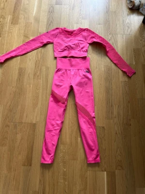 Rosa träningstights och crop top set - Säljer ett matchande träningsset i stark rosa färg med långärmad crop top och höga tights. Setet har meshdetaljer på armar och ben för extra ventilation och stil. Perfekt för gymmet eller yoga. Materialet är stretchigt och följsamt för maximal rörelsefrihet.