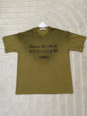 Olivgrön Acne Studios t-shirt - Olivgrön t-shirt från Acne Studios med svart text 'Acne Studios STOCKHOLM 1996' på bröstet. Klassisk passform med rund hals och korta ärmar. Perfekt för en avslappnad och trendig look.