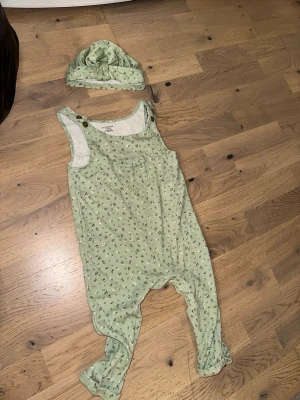 Grönt set med body och mössa - Sött set med ljusgrön ärmlös body och matchande mössa. Bodyn har små blommönster, bröstficka och knappar på axlarna. Mjukt och bekvämt material, perfekt för mysiga dagar hemma.Grönt set med body och mössa