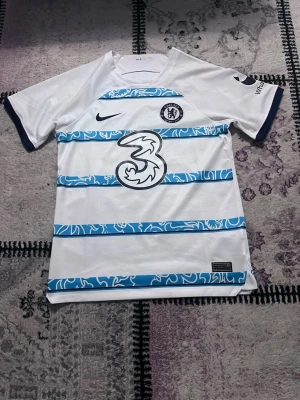 Chelsea vit och blå matchtröja Nike - Chelsea FC matchtröja i vitt med blåa grafiska ränder och klubbmärke på bröstet. Tröjan har korta ärmar med mörkblå kant och stor sponsorlogga på framsidan. Tillverkad i lätt och ventilerande material, perfekt för fotbollsträning eller match.