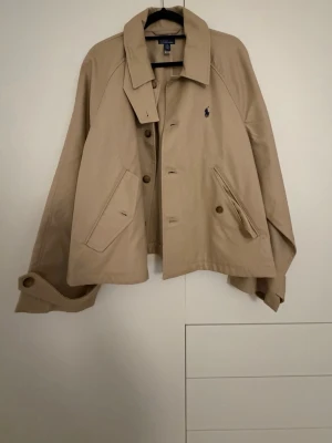 Beige trenchcoat från Polo Ralph Lauren - Klassisk beige trenchcoat från Polo Ralph Lauren med knäppning framtill, två snedställda fickor och justerbara ärmslut med knappar. Jackan har en rak passform och ikonisk logga broderad på bröstet. Perfekt för dig som gillar tidlös och stilren design.