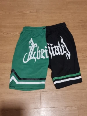 Gröna och svarta basketshorts - INSTABOX!!! Säljer ett par unika basketshorts i grönt och svart med vit och grön rand längs benen. Ena benet är grönt och det andra svart, med stor vit text framtill. Perfekta för dig som vill sticka ut på planen eller gatan.
