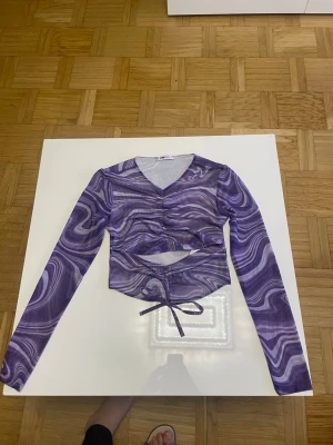 Lila mesh topp från FB Sister - Trendig lila mesh topp från FB Sister med abstrakt mönster, v-ringning och långa ärmar. Toppen har snörning i midjan och croppad passform som ger en edgy vibe. Perfekt för dig som vill sticka ut med coola detaljer och unik look.