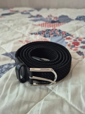 Grisch bälte  - Snyggt svart flätat bälte med ett stilrent silverfärgat metallspänne. Bältet har detaljer i svart skinn vid spännet och änden, vilket ger en klassisk look. Perfekt för att lyfta vilken outfit som helst.