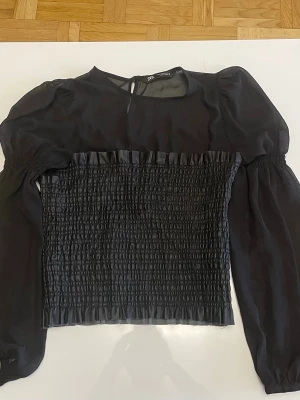 Svart blus med puffärm från Zara - Trendig svart blus från Zara med puffiga långärmar och smockad kropp. Blusen har rund halsringning och knäppning i nacken. Materialet är lätt och luftig polyester, perfekt för att skapa en chic look.