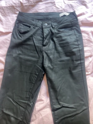 Svarta bootcut läderbyxor VERO MODA - Säljer ett par svarta läderbyxor som är bootcut och lågmidjade och klassisk femficksdesign. Byxorna stängs med knapp och dragkedja. Materialet har en lätt glansig finish som ger en cool look.