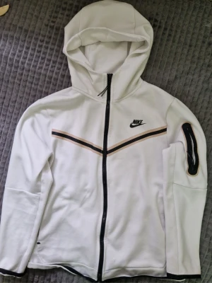 Vit Nike hoodie med dragkedja - Snygg vit hoodie från Nike med hel dragkedja framtill och svart Nike-logga på bröstet. Hoodien har en huva, svarta och bruna detaljer över bröstet samt en ficka med dragkedja på ena ärmen. Tillverkad i mjukt material som känns skönt mot huden.