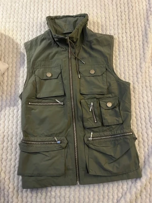 Grön väst Peak Performance XS - Snygg grön väst från Peak Performance i storlek XS. Västen har flera fickor med både dragkedja och knappar, hög krage med snörning och cool utility-look. Perfekt för dig som gillar funktionella detaljer och stilren design.