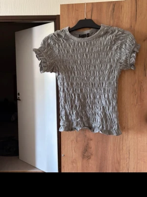 Grå ribbad topp med volang - Säljer en grå ribbad topp med korta ärmar och volangdetaljer vid ärmslut och nederkant. Toppen har en tight passform och ett stretchigt material som ger en snygg struktur. Perfekt att styla med jeans eller kjol.
