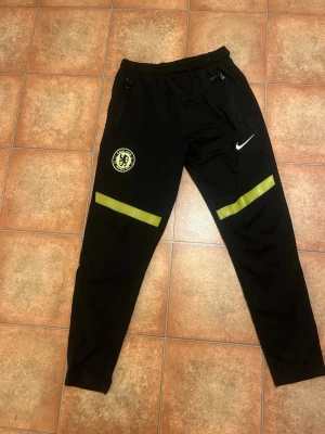 Chelsea FC svarta träningsbyxor Nike - Svarta träningsbyxor från Nike med Chelsea FC-logga på vänster ben och två gula reflexdetaljer ovanför knäna. Byxorna har dragsko i midjan och smal passform. Perfekta för fotbollsträning eller gymmet.