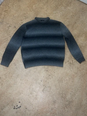 Mörkgrå stickad tröja från 157 - Mysig mörkgrå stickad tröja från 157 med rund halsringning och ribbade muddar vid ärmslut och nederkant. Perfekt för kyliga dagar och enkel att matcha med olika outfits. Materialet känns mjukt och värmande.