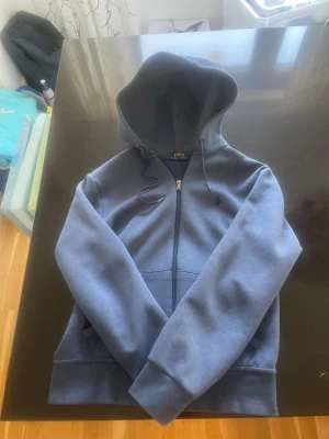 Blå zip hoodie från Polo Ralph Lauren - Snygg blå hoodie med dragkedja från Polo Ralph Lauren. Klassisk design med huva och dragsnören, två fickor framtill och diskret logga på bröstet. Perfekt för en chill och avslappnad stil. Tillverkad i mjukt sweatshirtmaterial.