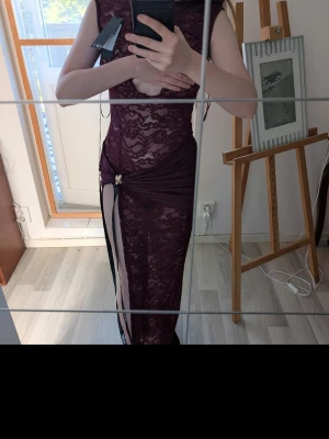 Vinröd spetsklänning med slits onyx dress - Vinröd långklänning i spets med ärmlös design och höga slitsar. Klänningen har dekorativa guldringar framtill och transparent spetsmaterial. Snygg och unik detalj med cut-out vid bysten och omlottkänsla vid höften. Lappar kvar, form sittande men inte obekvämt tight.