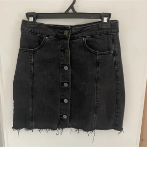 Svart jeanskjol med knappar - Snygg svart jeanskjol med knappar framtill och rå kant nertill. Kjolen har två fickor fram och en klassisk denimlook.